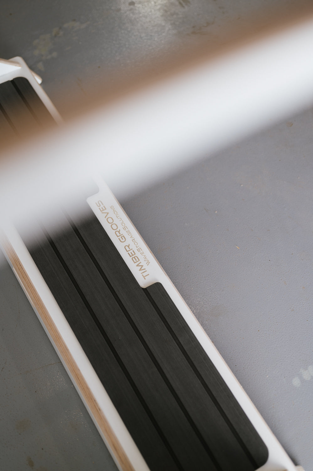 A close-up of a gray and white object labeled "Timber Grooves Surfboard Rack Freestanding" laying flat on a gray floor surface, perfect for those interested in custom surf racks.