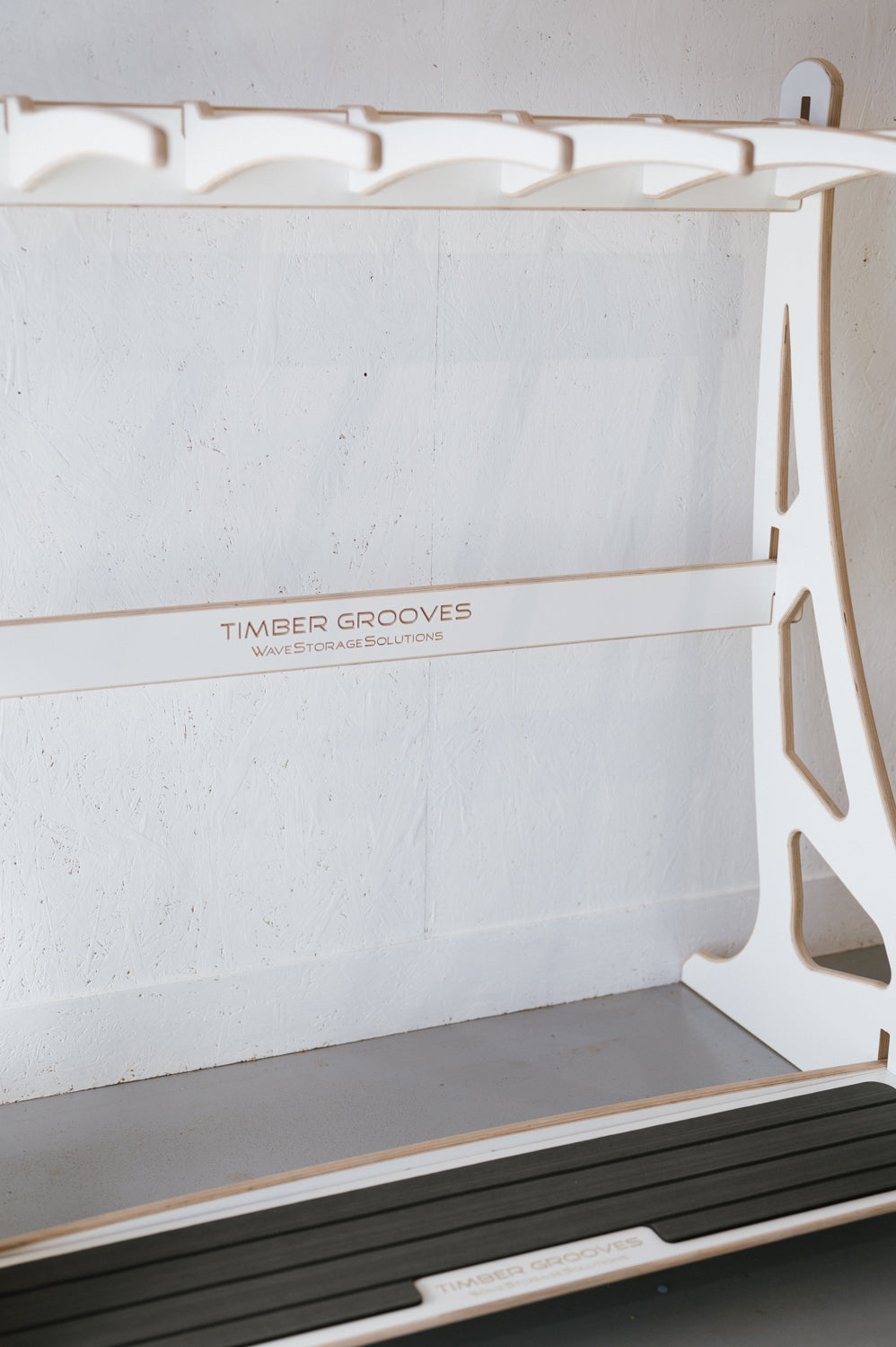 A white wooden rack labeled "Timber Grooves Surfboard Rack Freestanding" stands against a white textured wall, designed for organizing and storing surfboards. This custom surf rack combines practicality with effortless style.