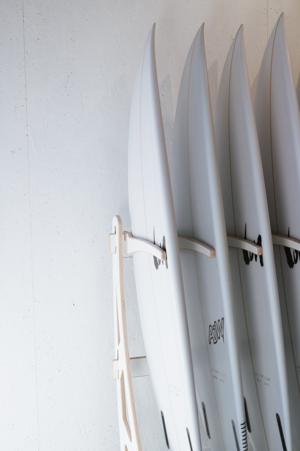 Four white surfboards are neatly arranged upright on a wooden rack against a white wall. Wholesale inquiries for our custom Surfboard Rack Freestanding by Timber Grooves can help you organize your gear like a pro, ensuring everything is stylishly displayed and easily accessible.