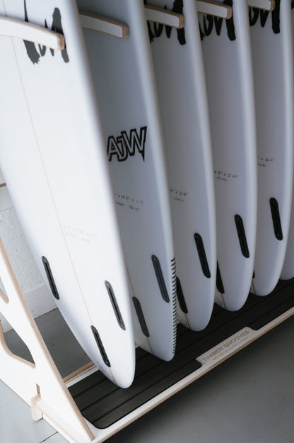 A row of white surfboards with black logos stands vertically in a wooden rack, ideal for wall-mounted racks at surf shops. The base of the Surfboard Rack Freestanding reads "Timber Grooves.