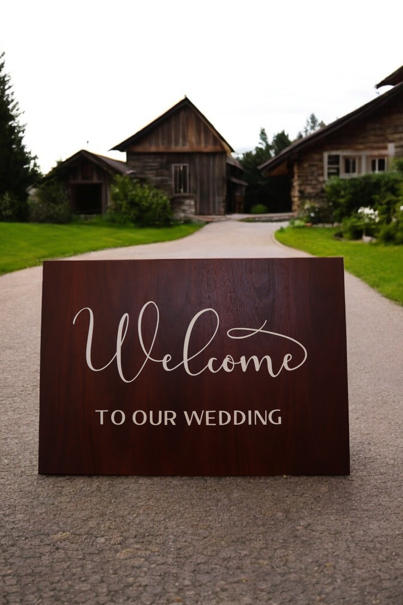 A rustic elegant atmosphere enhances the path, adorned with a Personalised Timber Wedding Welcome Sign by Timber Grooves that warmly welcomes guests. Nestled among lush greenery, this custom-engraved creation directs them to charming rustic buildings, perfectly setting the mood for the celebration.