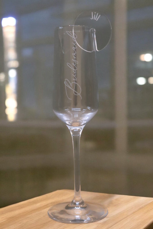 A champagne flute with "Bridesmaid" custom-engraved on it sits elegantly on a wooden surface, adorned with a Timber Grooves Personalised Acrylic Drink Tag labeled "Jill.