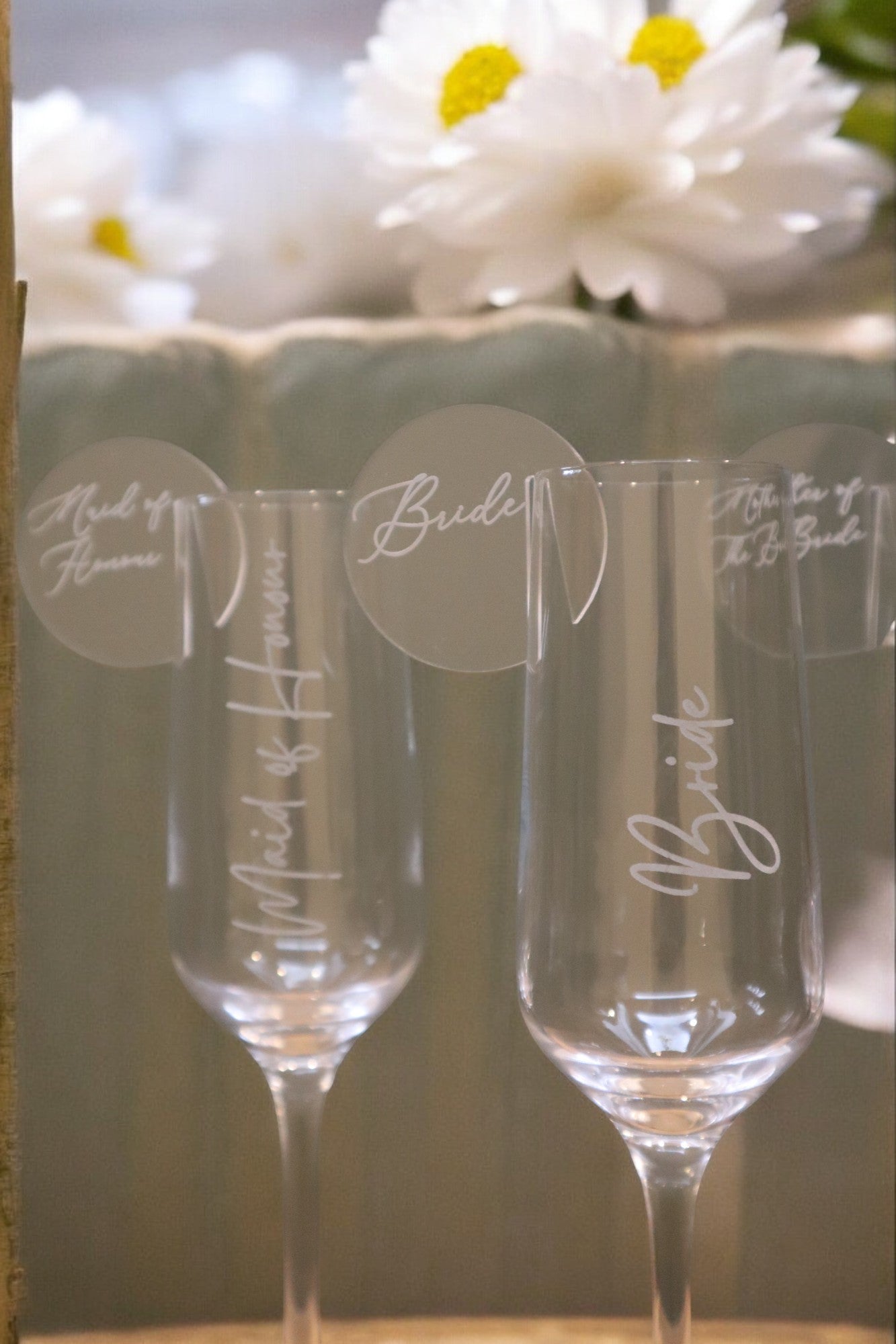 A pair of Personalised Champagne Flutes by Timber Grooves, each with circular tags inscribed "Maid of Honor" and "Bride," beautifully set against a floral backdrop.