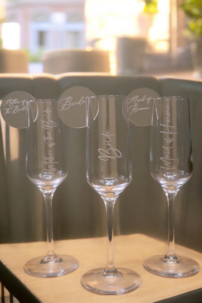 Three champagne flutes labeled "Bride," "Maid of Honor," and "Mother of the Bride" rest on an elegant table setting, adorned with sunlight. Each glass features a Personalised Acrylic Drink Tag from Timber Grooves, adding a personal touch to the celebration.