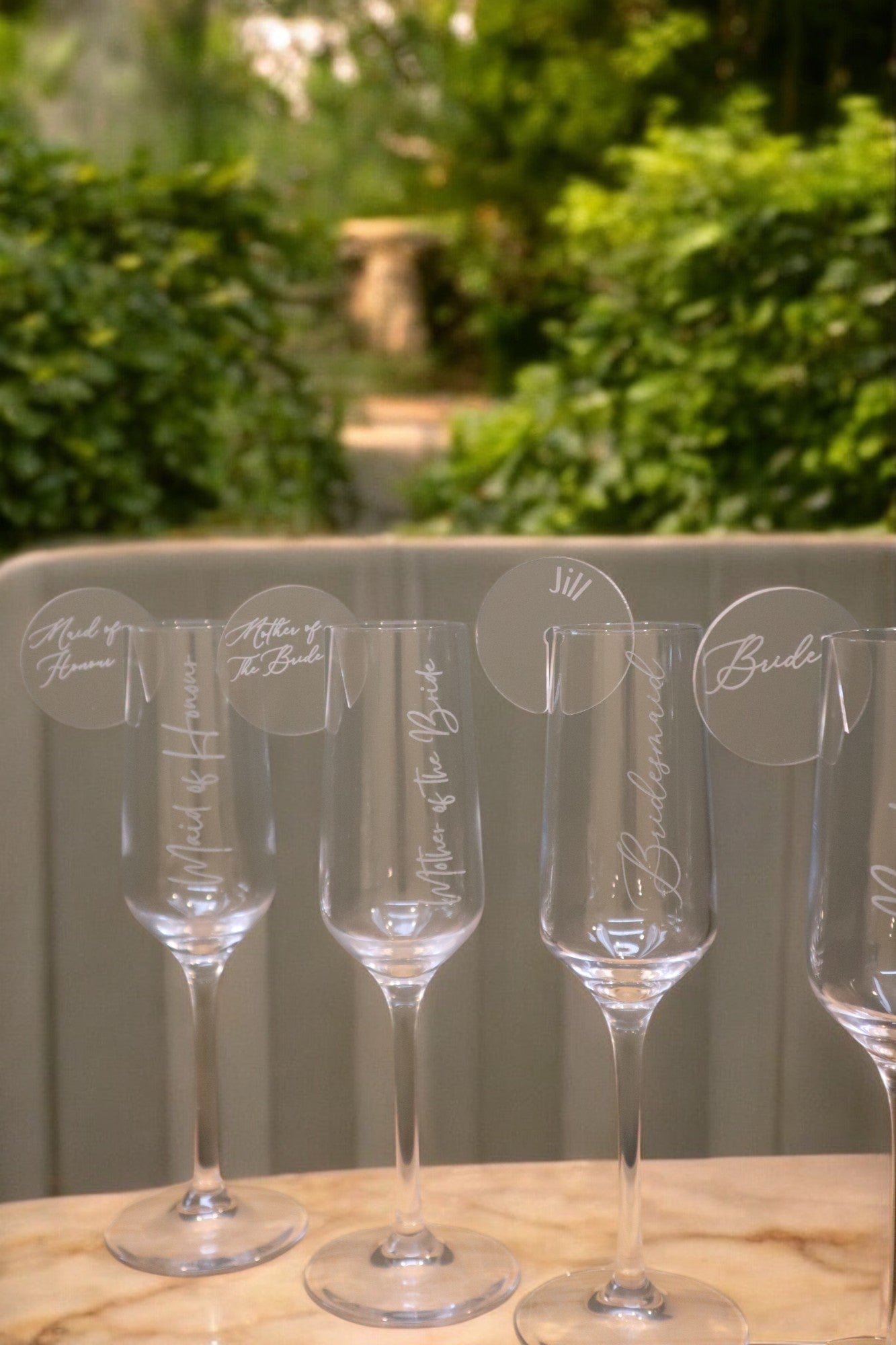 A set of four Timber Grooves Personalised Champagne Flutes, each engraved with names and wedding-related titles, are elegantly arranged on a table with greenery visible in the background.