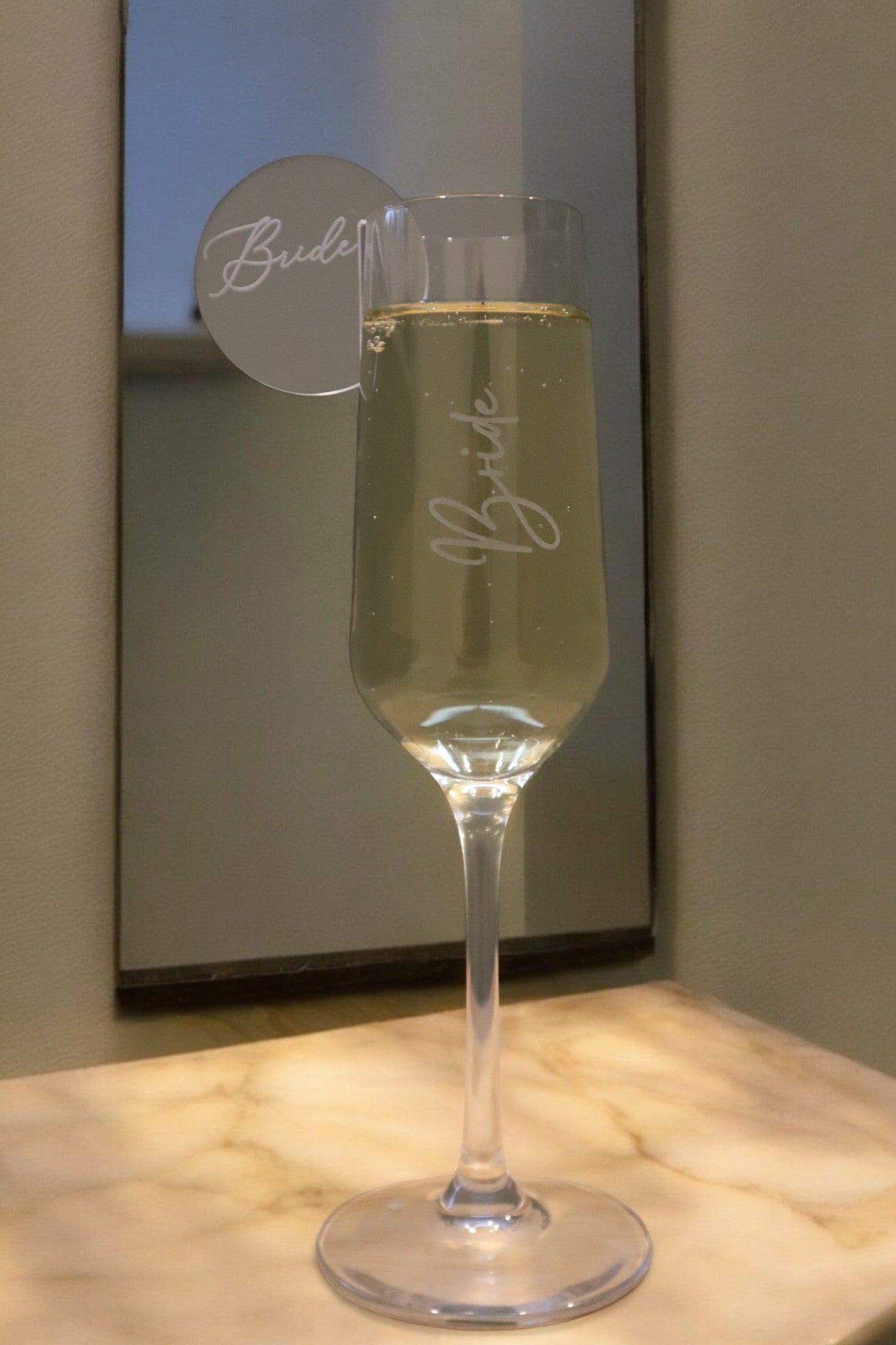 A champagne flute from Timber Grooves, personalized with the label "Bride" and featuring a matching tag, sits on a marble surface in front of a mirror.