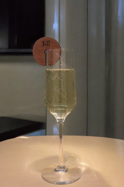 A champagne flute with "Brut" etched on it rests elegantly on a table, holding a pale bubbly drink. A Personalised Timber Drink Tag from Timber Grooves, custom-engraved with "Jill," graces the glass rim, adding a special touch perfect for any wedding or celebration.