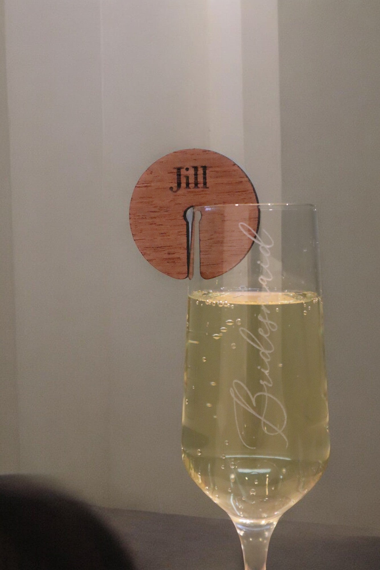 A champagne flute with "Bridesmaid" etched on it stands elegantly in the foreground. Behind it, a wooden coaster featuring a custom-engraved tag from Timber Grooves, known as the Personalised Timber Drink Tag and engraved with the name "Jill," is mounted on the wall, perfect for adding that personalized touch to any wedding or celebration.