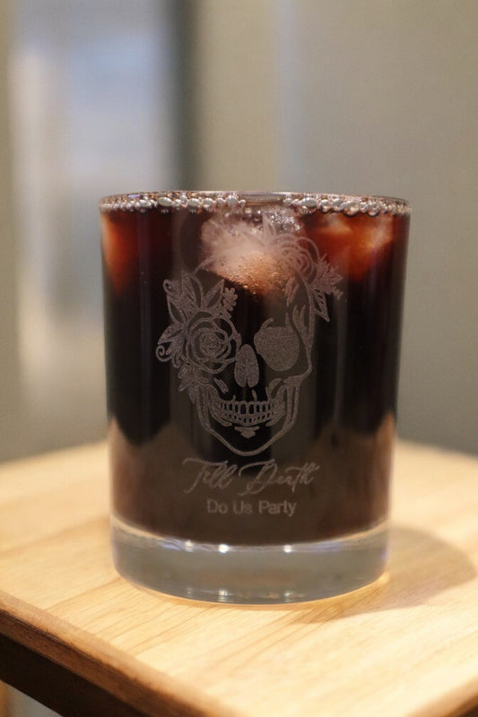 An Engraved Whiskey Glass by Timber Grooves, featuring a skull and floral design with the inscription "Till Death Do Us Party," resting on a wooden surface filled with dark iced coffee.