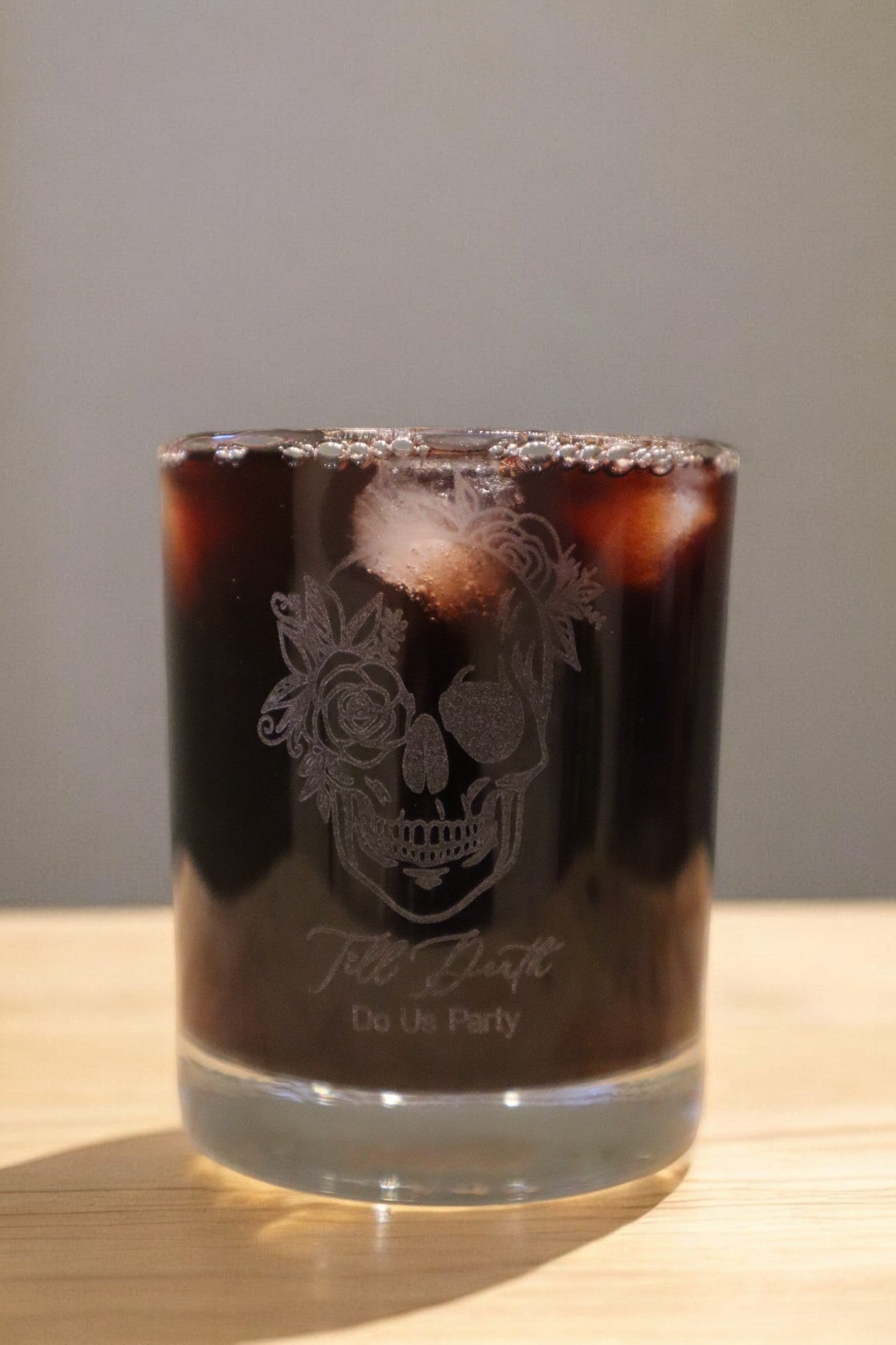 A Timber Grooves Engraved Whiskey Glass, featuring a dark iced beverage and adorned with a decorative skull design along with the phrase "Till Death Do Us Party," rests on a wooden surface.