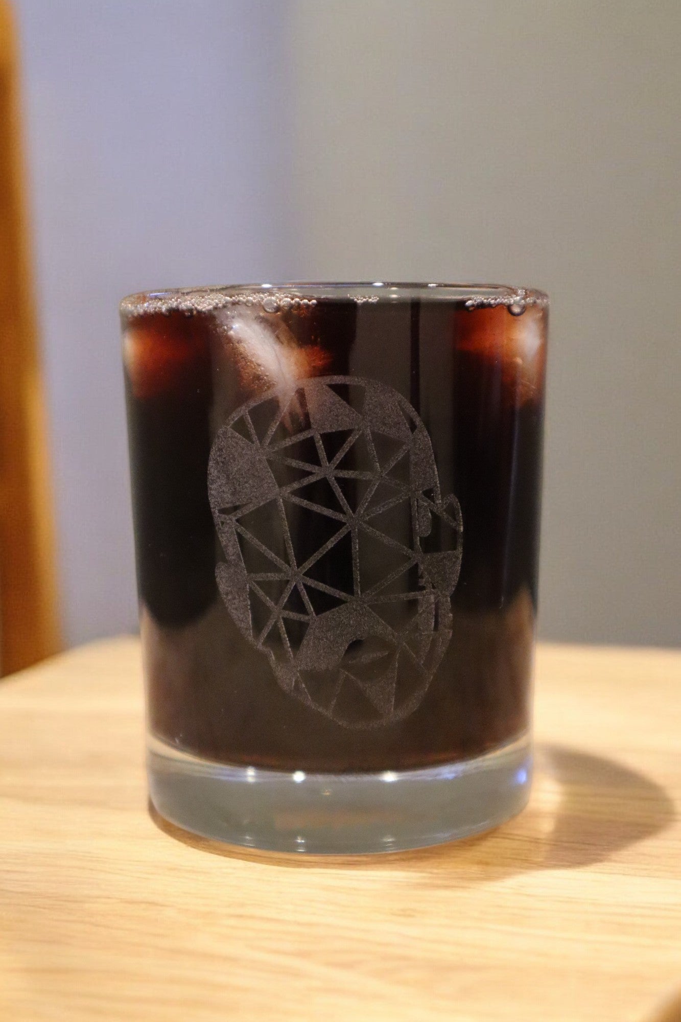 A glass of dark iced beverage in an Engraved Whiskey Glass by Timber Grooves, featuring a geometric skull design, sits on a wooden surface.