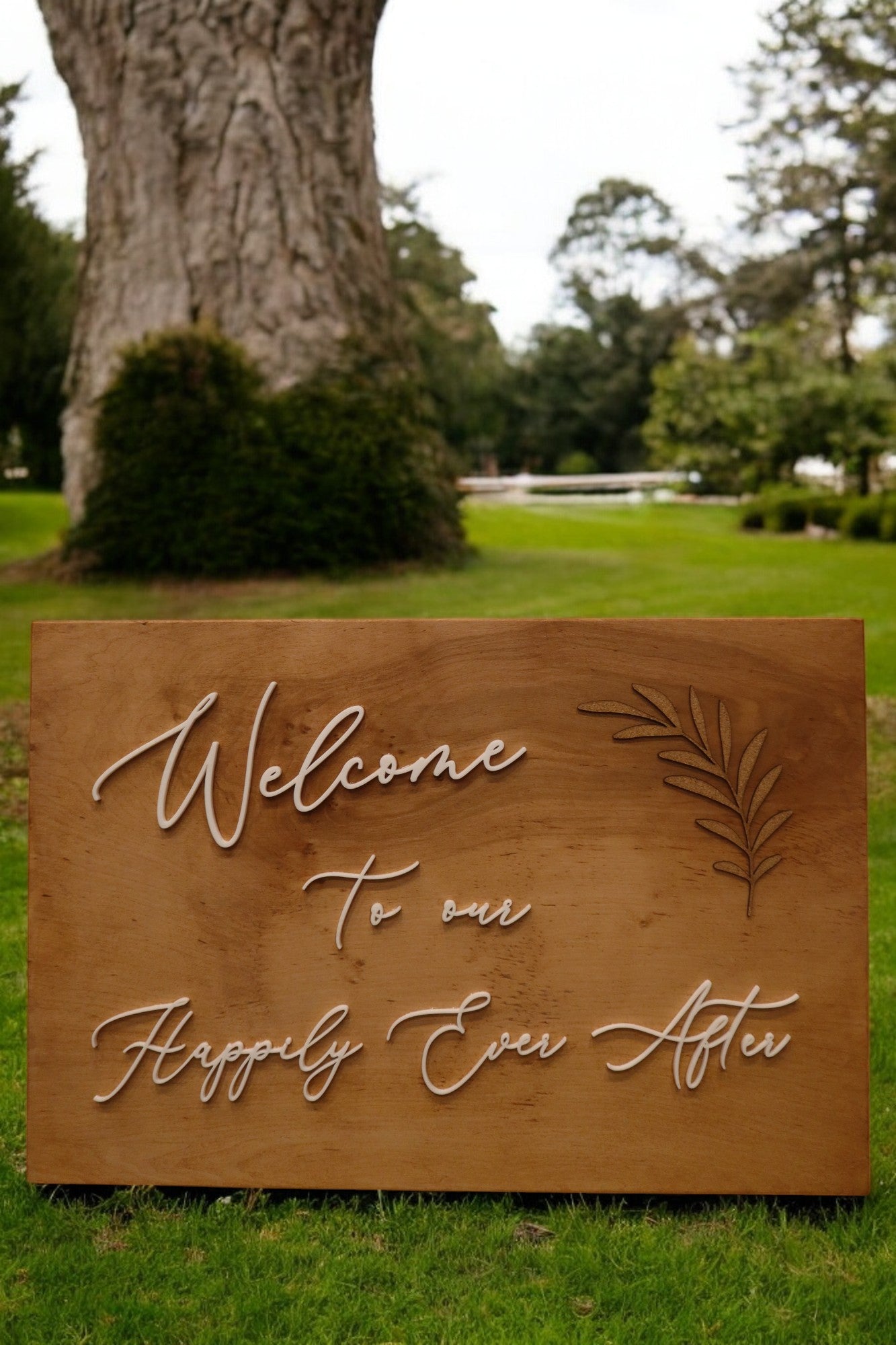 The charming Personalised Timber Wedding Sign from Timber Grooves features the phrase "Welcome to our Happily Ever After," elegantly embellished with decorative leaf details. This delightful wedding keepsake is beautifully displayed on the grass, surrounded by trees, adding a personal touch to your special day.