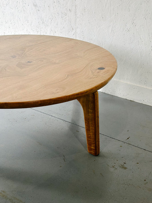 A round wooden Marri Coffee Table by Timber Grooves with three curved legs, meticulously handcrafted from Australian Hardwood, placed on a gray concrete floor against a textured white wall.