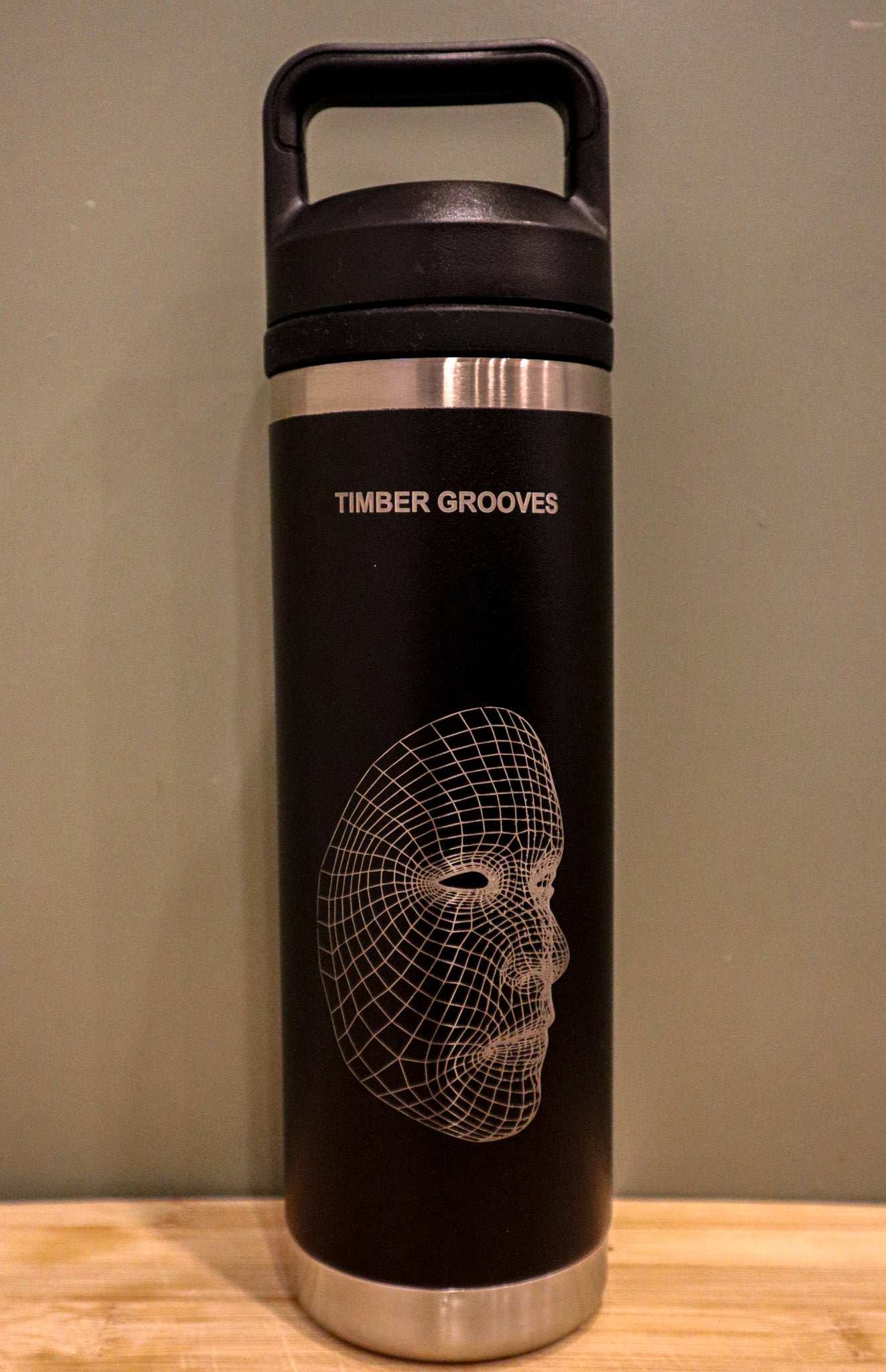 Black and stainless steel tumbler from Timber Grooves featuring a wireframe face design, similar to a custom laser-engraved water bottle, resting on a wooden surface.