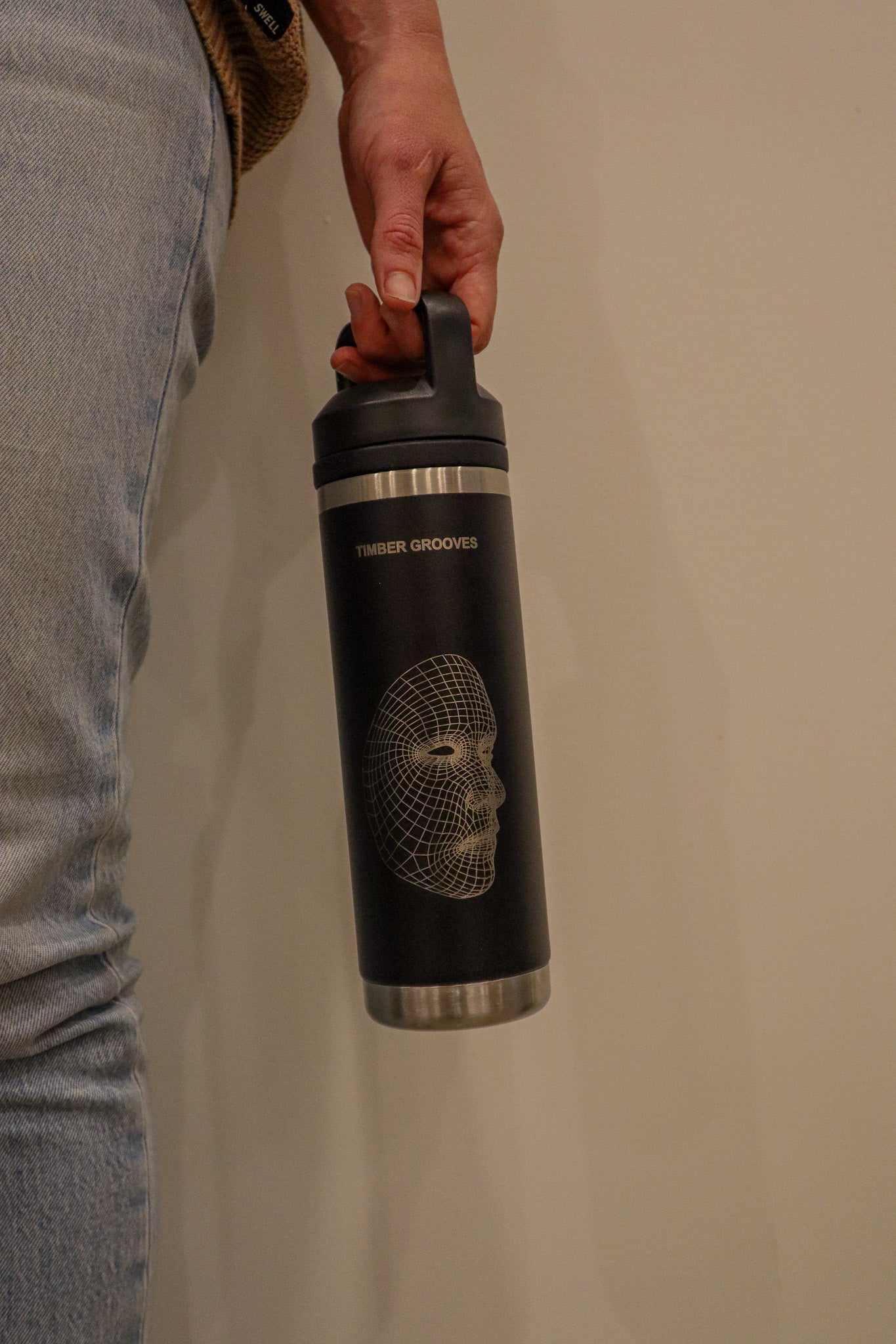 A person holds a Personalised Laser Engraved Water Bottle from Timber Grooves, showcasing a geometric skull design and "Timber Grooves" text on its black insulated exterior.