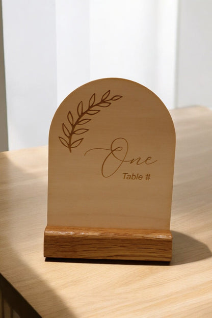 The Engraved Timber Wedding Table Number by Timber Grooves features the word "One" with a decorative leaf design, adding rustic charm to any wedding decor. This personalized piece elegantly stands on the table.