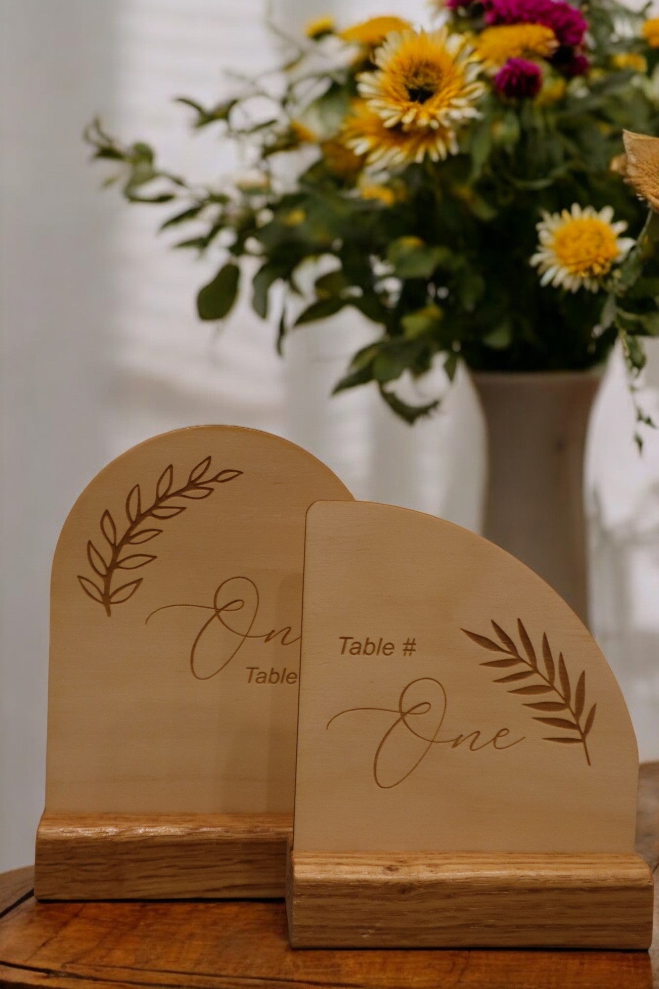 The Engraved Timber Wedding Table Number by Timber Grooves, featuring intricate leaf designs and engraved with "Table # One," exudes rustic charm on a wooden surface. Behind them, a vase filled with colorful flowers enhances the enchanting ambiance of this personalized decor.