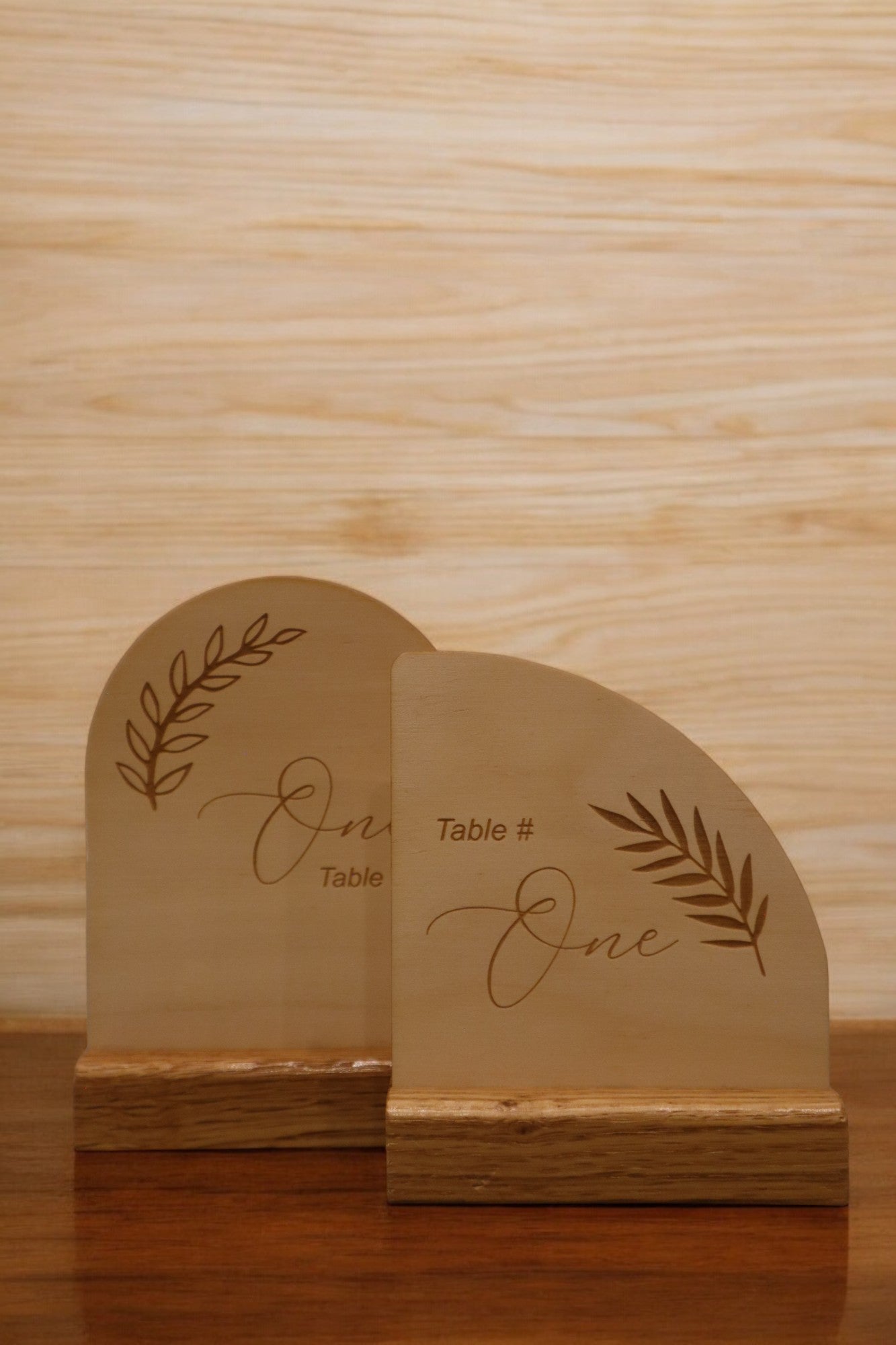 Two Engraved Timber Wedding Table Numbers by Timber Grooves exude rustic charm with engraved text "Table # One" and decorative leaf designs, making them perfect for personalized wedding decor. Resting elegantly on a wooden surface, these pieces add a touch of natural beauty to any celebration.