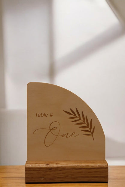 The Engraved Timber Wedding Table Number from Timber Grooves showcases rustic elegance with "Table # One" beautifully engraved alongside a carved leaf design. It stands gracefully on a wooden base, making it perfect for personalized decor at weddings or intimate gatherings.