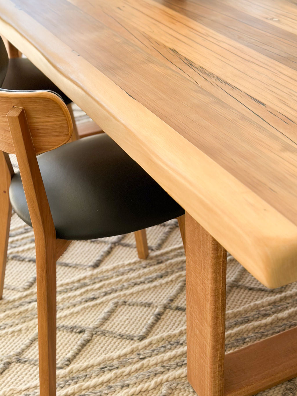 A close-up view of a handcrafted dining table with a matching wooden chair that has a black cushioned seat, all set upon a patterned rug. The Timber Grooves Farmhouse Dining Table adds rustic charm to the space.