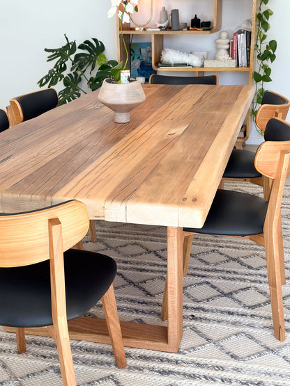 A handcrafted Farmhouse Dining Table from Timber Grooves with a Blackbutt table base is surrounded by six wooden chairs with black seats. A decorative vase sits on the table, a patterned rug is underneath, and a shelf with various items is in the background.