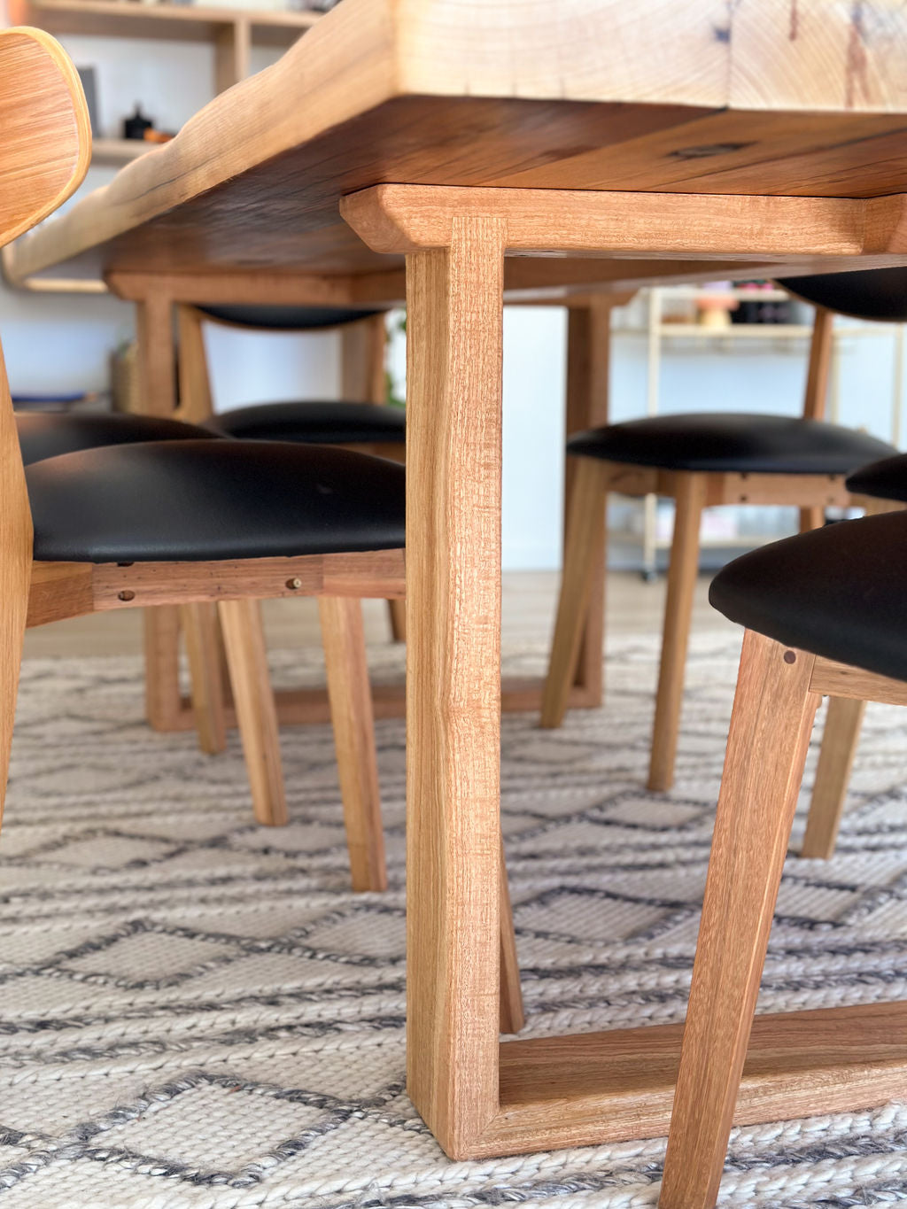 Close-up view of a modern **Timber Grooves Farmhouse Dining Table** with matching chairs, featuring black cushioned seats, placed on a patterned woven rug. The handcrafted dining table boasts a Blackbutt table base that adds an elegant charm to the ensemble.