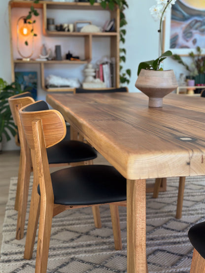 A Farmhouse Dining Table by Timber Grooves with a Blackbutt table base holds a potted plant centerpiece, surrounded by wooden chairs with black seats. The background features a bookshelf adorned with decor and plants.