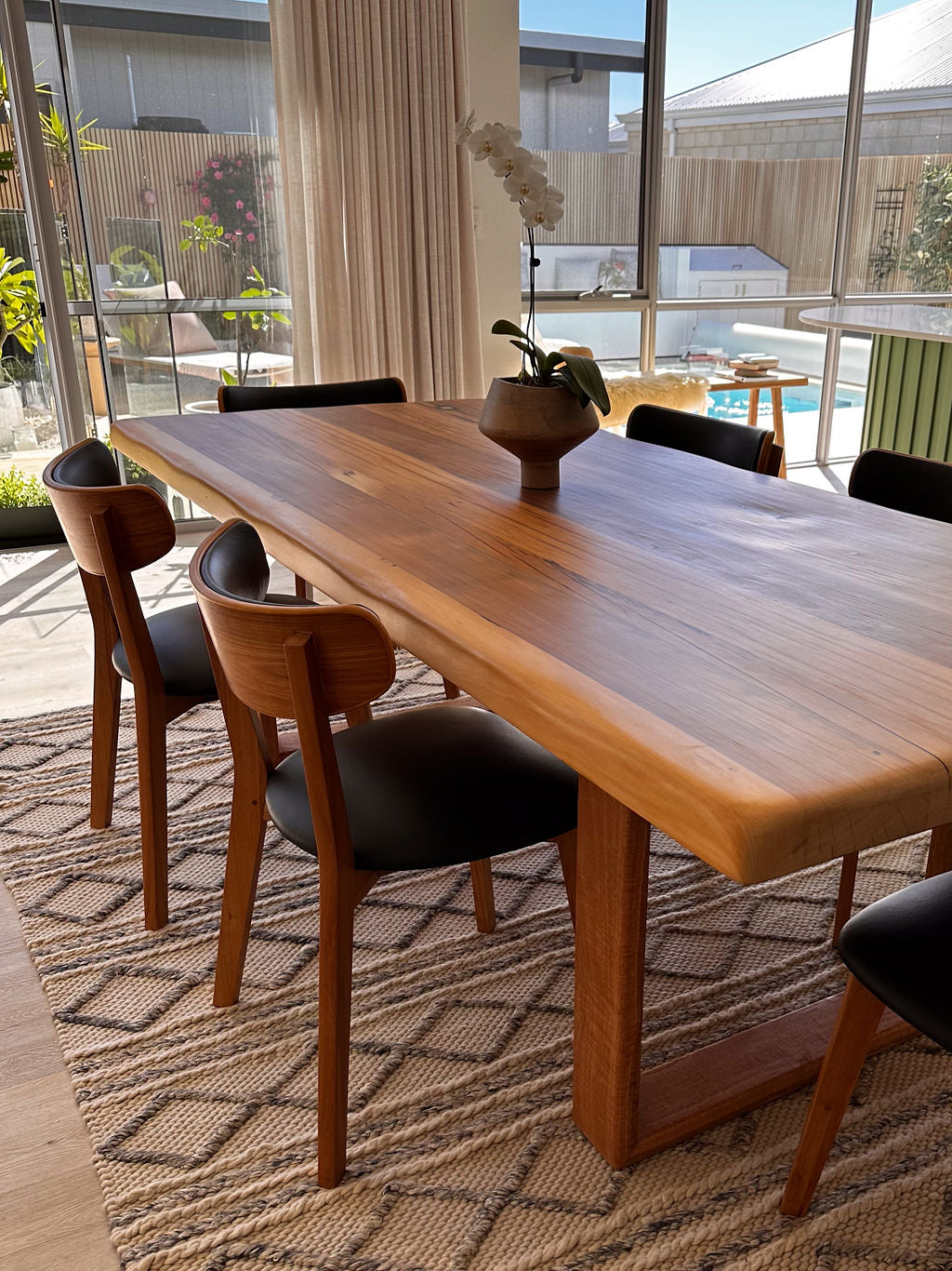 A Timber Grooves Farmhouse Dining Table stands adorned with black chairs on a woven rug in a bright room. Large windows offer a view of an outdoor pool, while a small potted plant graces the center of the table.