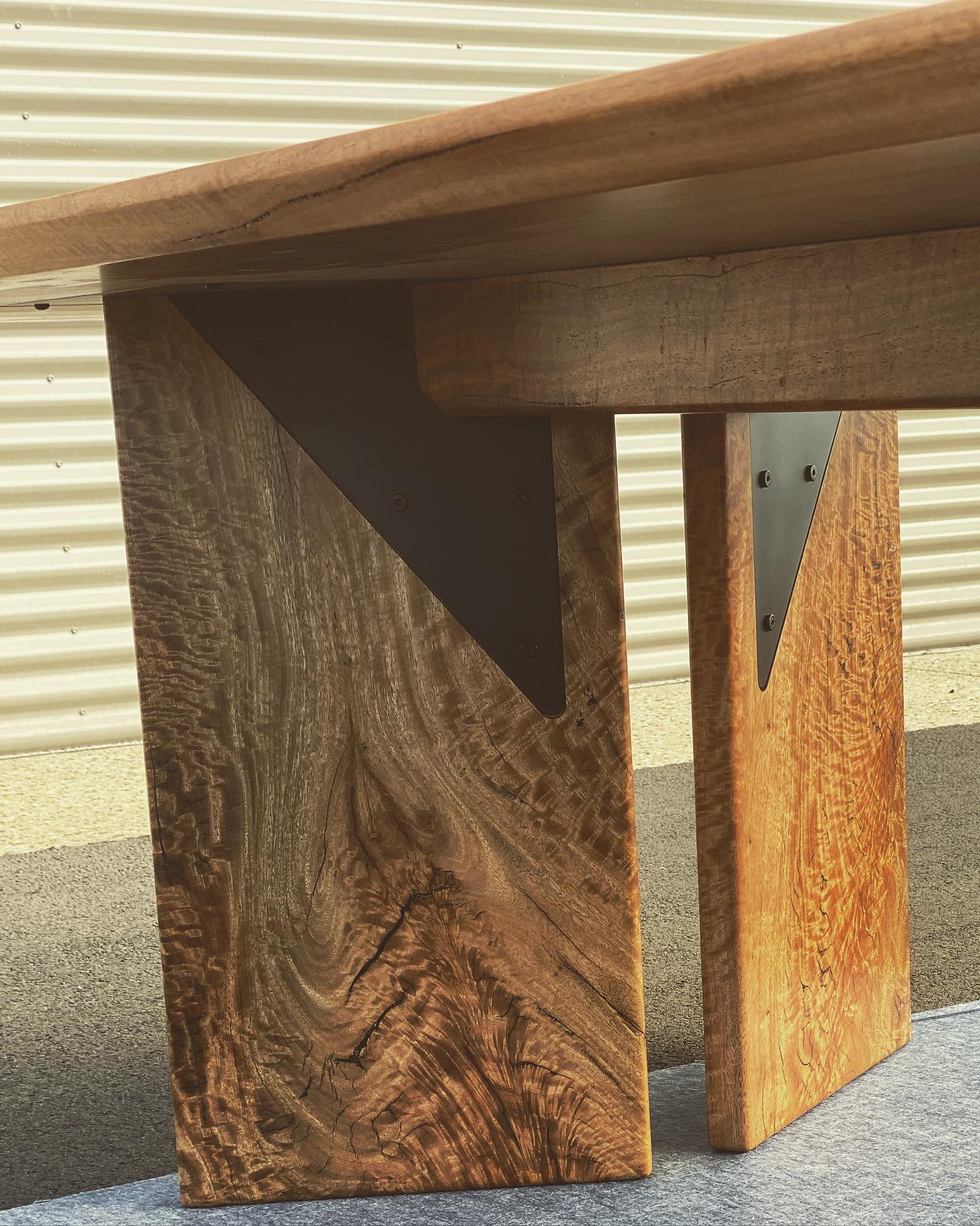 Close-up view of a handcrafted Timber Grooves Marri Dining Table with thick legs featuring black metal brackets, positioned on a carpeted surface in front of a corrugated metal wall, highlighting its exquisite 3D grain.