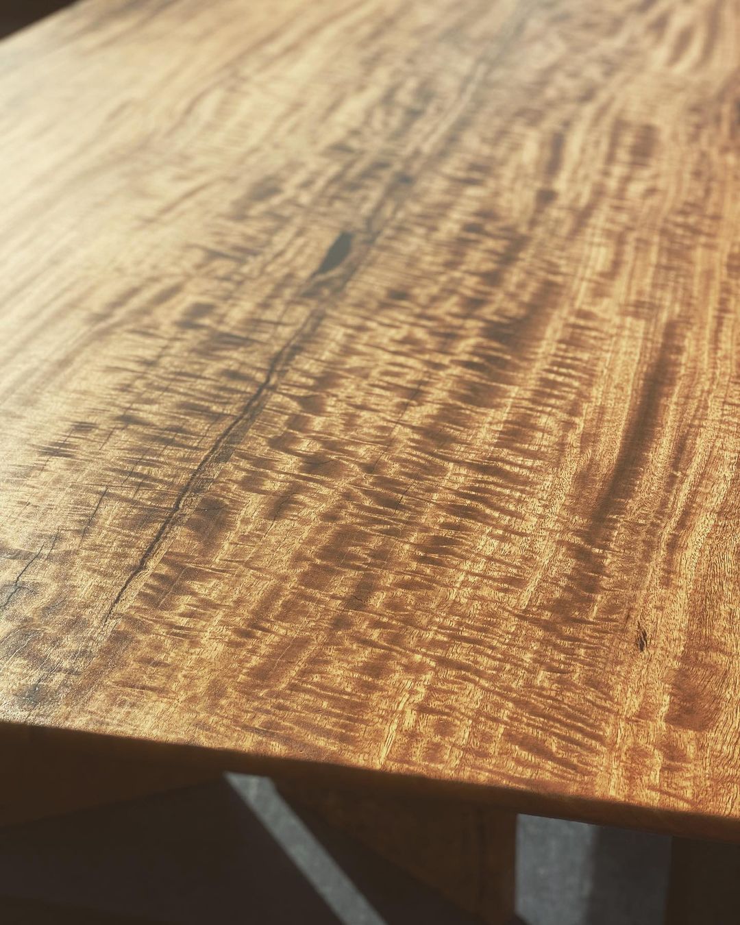 Close-up of a handcrafted Timber Grooves Marri Dining Table's surface displaying a wavy, 3D grain texture with natural variations. The wood appears polished and reflects light, highlighting its intricate pattern and warm hue.